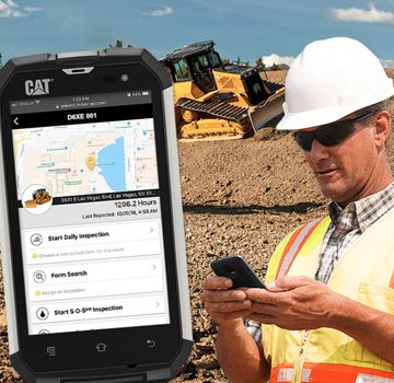 A person holding a handheld device on displaying the Cat app with a Cat dozer behind him